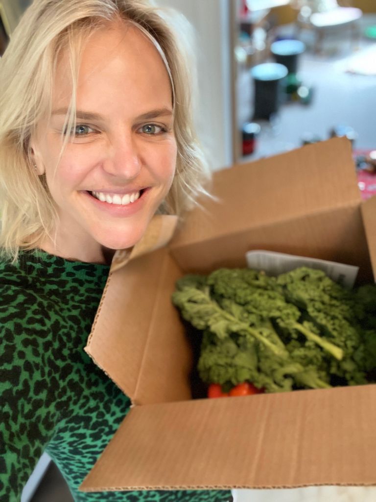A woman with blond hair, smiling, holding a box of fresh vegetables including broccoli and tomatoes from Rubies In The Rubble.