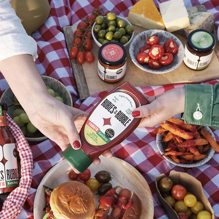 A person pours Rubies In The Rubble ketchup on a burger at a picnic with various foods like cheese, olives, and chips arranged around on a red checkered cloth.