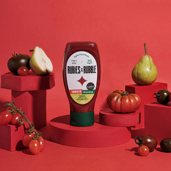 A bottle of Rubies in the Rubble tomato ketchup on a red background adorned with tomatoes, pears, and display stands.