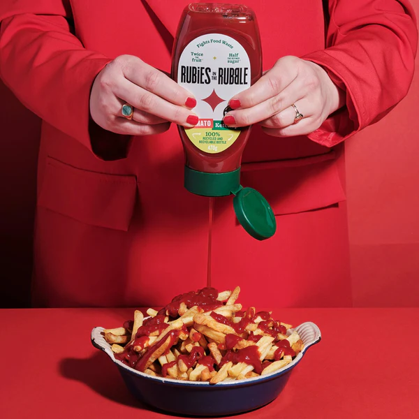 A person in a red jacket pours Rubies In The Rubble ketchup from a bottle onto a bowl of french fries, set against a red background.