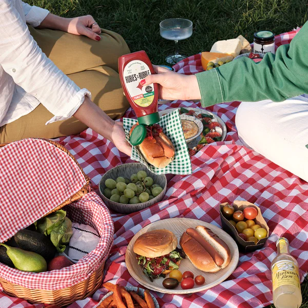 Two people enjoying a picnic with hot dogs and various snacks on a red and white checkered blanket, one person pouring Rubies In The Rubble ketchup.