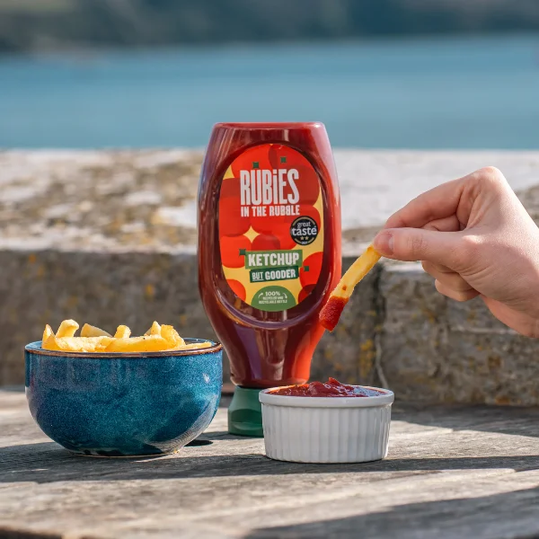 Image of Rubies in the Rubble ketchup with a bowl of chips and a ramekin full of ketchup. A hand is dipping the chip into the ketchup. The background is at the sea side.