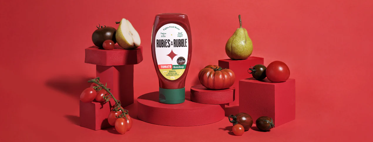A bottle of tomato ketchup from Rubies in the Rubble surrounded by fresh vegetables and fruits on red pedestals against a red background.