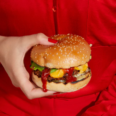 A hand holding a burger featuring Rubies In The Rubble ketchup, with lettuce, tomato, and cheese on a sesame seed bun against a red background.