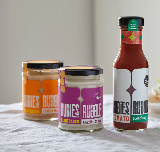 Three jars of Rubies In The Rubble condiments, including chili mayo, garlic mayo, and tomato ketchup, on a kitchen counter.