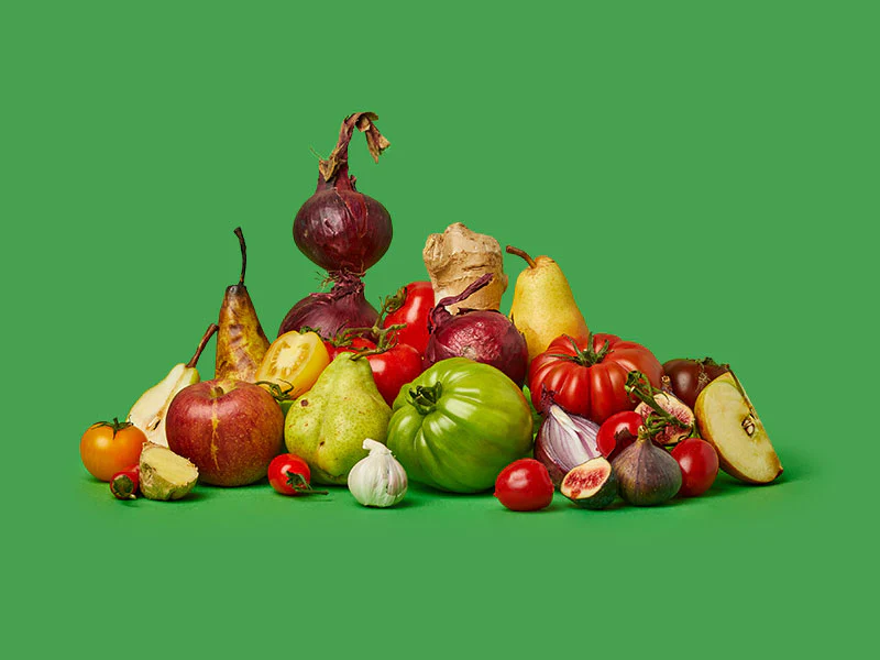 A variety of fresh produce, including tomatoes, pears, onions, and figs from Rubies In The Rubble, arranged on a green background.