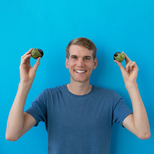 A smiling man in a blue t-shirt holding up two halves of an avocado against a blue background, branded as 