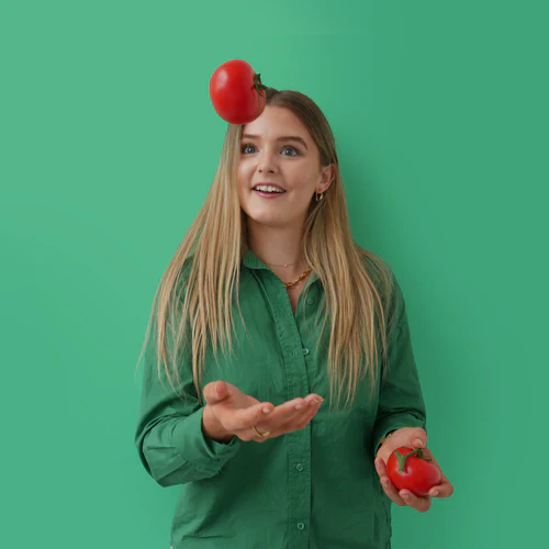 A young woman in a green shirt tosses a red tomato up in the air, balancing another on her head, against a green background, evoking the playful essence of 