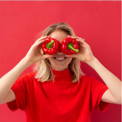 Woman in red shirt holding two red bell peppers over her eyes against a ruby background.