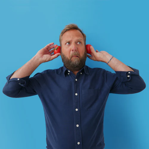 Man in blue shirt holding two Rubies In The Rubble apples to his ears, looking puzzled, against a blue background.