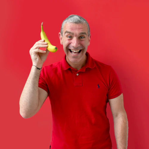 A smiling middle-aged man in a red shirt holding a banana like a telephone against a background of rubies in the rubble.