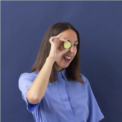 A woman in a blue shirt holds a slice of cucumber over her eye and smiles, standing against a plain blue background with the logo 