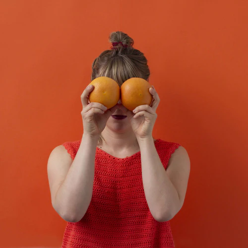 Woman in red dress holding two oranges over her eyes against a backdrop titled 