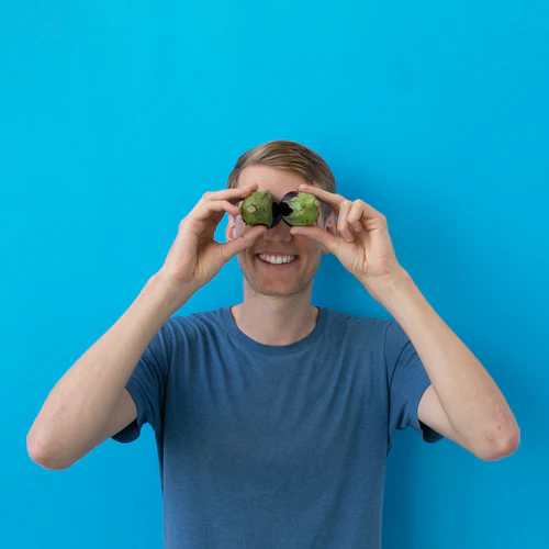 Man smiling and holding two avocado halves over his eyes against a blue background titled 
