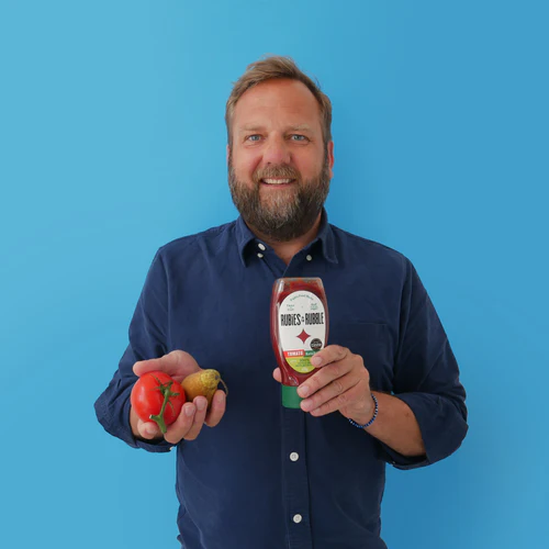 A smiling bearded man holding fresh vegetables in one hand and a bottle of Rubies In The Rubble's miracle sauce in the other, against a blue background.