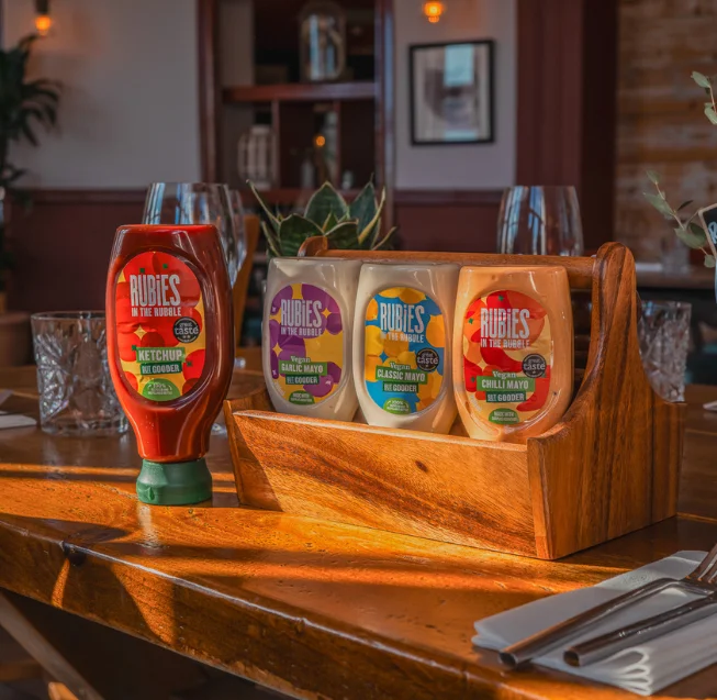 The Rubies in the Rubble Squeezy range of ketchup, garlic mayo, classic mayo and chilli mayo in a caddy on a tabletop in a pub setting.